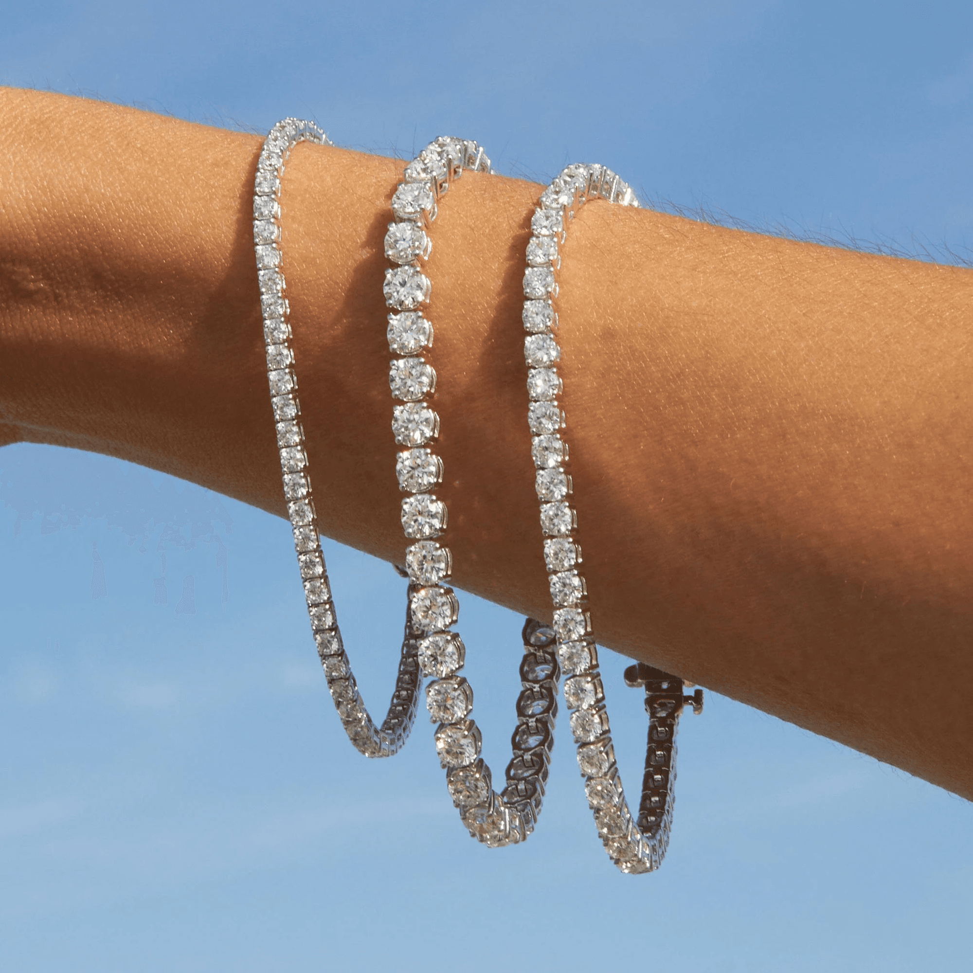 Three diamond tennis bracelets layered on a wrist against a blue sky